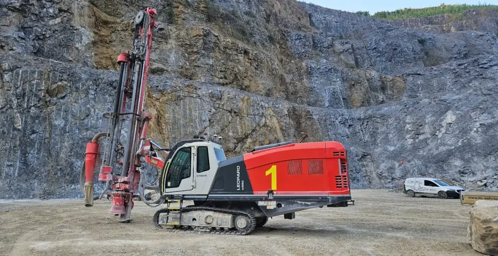 Red crawler drill rig beside a large rock in a terraced quarry.