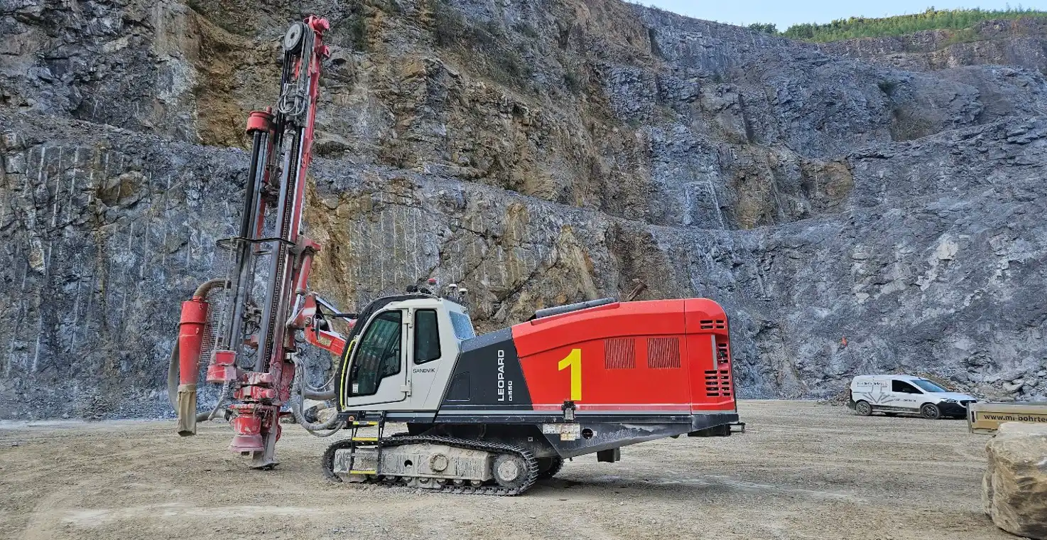 Red crawler drill rig beside a large rock in a terraced quarry.