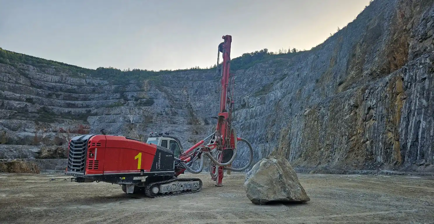 Red crawler drill rig beside a large rock in a terraced quarry.