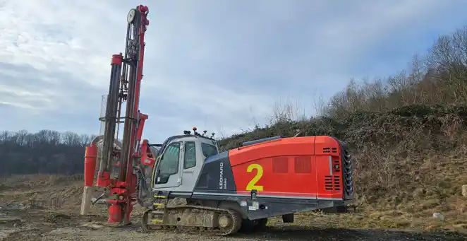 Red crawler drill rig beside a large rock in a terraced quarry.