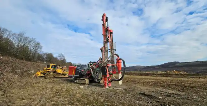 Red crawler drill rig with a raised mast on a large earthworks site.